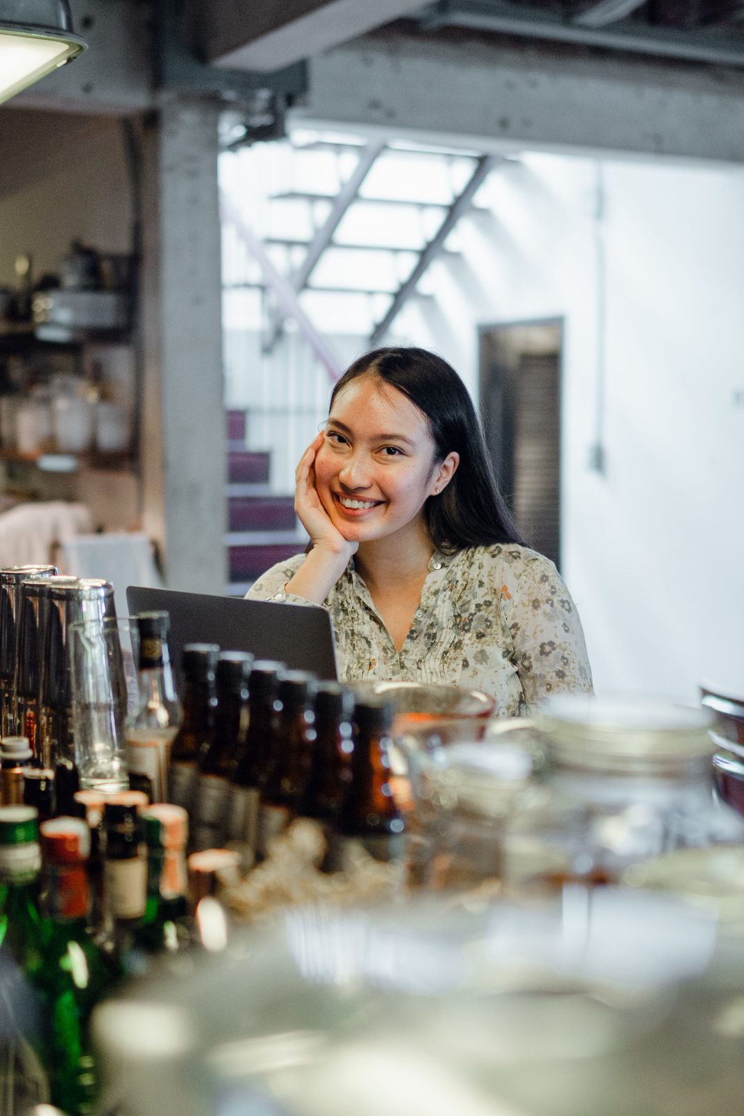 A shop owner sits thoughtfully at her counter, resting her chin on one hand beside a laptop.